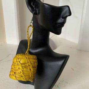 May include: A single earring featuring a bright yellow, woven straw cube suspended from a braided straw loop. The earring is attached to a silver-toned metal hook finding, showcasing a textured, natural fibre construction in a vibrant, solid yellow hue.