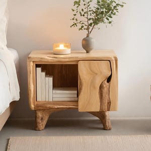 Puede incluir: Una mesita de noche de madera natural con un diseño de borde vivo. La mesita de noche presenta un pequeño armario con una puerta tallada única, una estantería con libros y una vela encendida. Una pequeña vasija con vegetación encima.