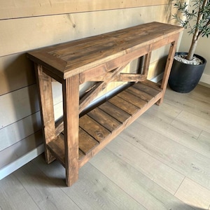 Könnte beinhalten: Ein rustikaler Konsolentisch aus Holz mit natürlichem Finish. Der Tisch hat eine lange, rechteckige Oberseite, ein unteres Regal und eine X-förmige Stütze an den Seiten. Der Tisch ist ca. 152 cm lang, 38 cm breit und 76 cm hoch.