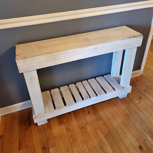 May include: A rustic, light-coloured wooden console table with a distressed finish. The table features a long, rectangular top, supported by thick, painted legs and a slatted shelf. The table is approximately 152 cm long.