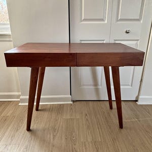 Op de afbeelding: Een houten bureau in mid-century modern stijl met een rechthoekig blad en vier taps toelopende poten. Het bureau heeft een warme bruine kleur en lijkt van massief hout te zijn. Het bureau is ongeveer 122 cm breed en 61 cm diep.
