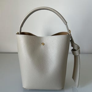 May include: A pale grey bucket bag with a rounded handle and a long strap detail. The bag has a textured surface and a small gold-coloured button closure. The interior is a lighter shade of brown.