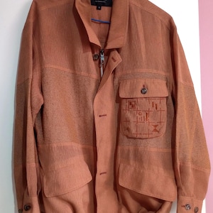 Könnte beinhalten: Eine terrakottafarbene Jacke mit durchgehendem Reißverschluss, Knopfleiste und einer Brusttasche mit gestickten geometrischen Mustern. Das Kleidungsstück verfügt über einen strukturierten Netzeinsatz an Brust und Ärmeln, zwei untere Taschen und einen Kordelzug am Saum.