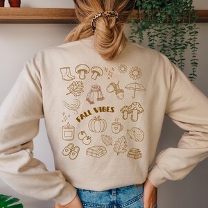 Könnte beinhalten: Ein beigefarbenes Sweatshirt mit Rundhalsausschnitt und einer braunen Strichgrafik auf der Rückseite. Das Design zeigt verschiedene herbstliche Symbole wie Pilze, einen Kürbis, einen Schal, Blätter, einen Regenschirm, eine Kerze und den Schriftzug FALL VIBES in Braun.