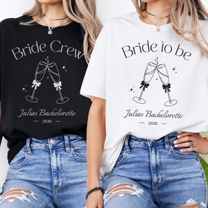 Könnte beinhalten: Ein schwarzes T-Shirt mit weißem Text Bride Crew und ein weißes T-Shirt mit schwarzem Text Bride to be. Beide T-Shirts zeigen eine Grafik von zwei anstoßenden Sektgläsern, den Text Julias Bachelorette und das Jahr 2026.