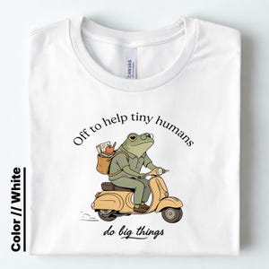 May include: A white t-shirt featuring a graphic of a green frog wearing glasses and a backpack, riding a tan scooter. The text above the frog reads "Off to help tiny humans" and the text below the frog reads "do big things" in black lettering.