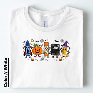 May include: A white t-shirt featuring a graphic of five anthropomorphic school supplies dressed in Halloween costumes: a blue crayon vampire, an orange pumpkin, a mummy glue stick, a black notebook, and a yellow pencil witch with a broom.