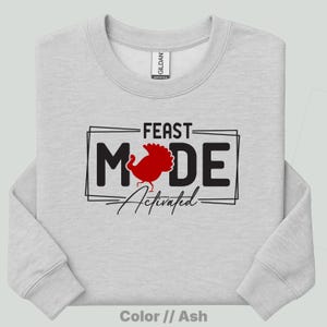 May include: An ash gray crewneck sweatshirt featuring a black graphic design with the text FEAST MODE ACTIVATED. The letter O in MODE is replaced by a red turkey silhouette. The text is enclosed in a black rectangular frame on the chest.