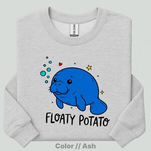 May include: An ash gray crewneck sweatshirt featuring a graphic of a bright blue manatee surrounded by small teal bubbles, a tiny red heart, and yellow stars. Below the manatee, the text reads "FLOATY POTATO" in a bold, black, hand-lettered font.