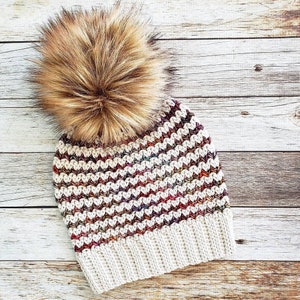 May include: A white crocheted beanie with a colorful chevron pattern and a brown faux fur pom pom.