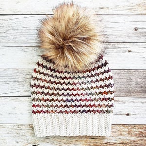 May include: A white crocheted beanie with a brown faux fur pom pom. The beanie has a striped pattern with shades of red, brown, and cream.