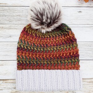 May include: A knitted winter hat with a gray ribbed brim and a colorful woven pattern in shades of orange, green, and red. The hat is topped with a fluffy, faux fur pom-pom in brown and white.