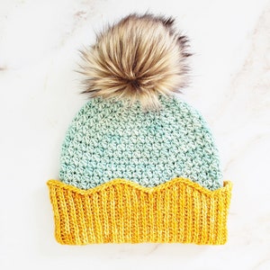 May include: A crocheted hat with a scalloped brim in a light blue and yellow color combination. The hat has a fluffy brown pom pom on top.