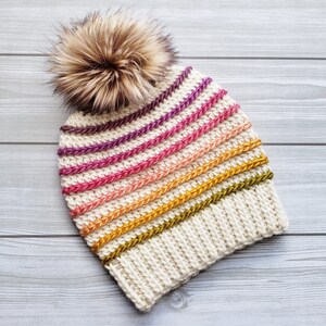Pode incluir: Um gorro branco de croch&ecirc; com um padr&atilde;o listrado em tons de rosa, laranja, amarelo e verde. O gorro tem um pompom marrom felpudo no topo.