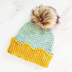 May include: A crocheted hat with a scalloped brim in teal and yellow with a brown faux fur pom pom.