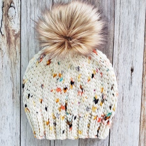 May include: A white knitted beanie with a colourful flecked pattern and a fluffy brown pom pom.