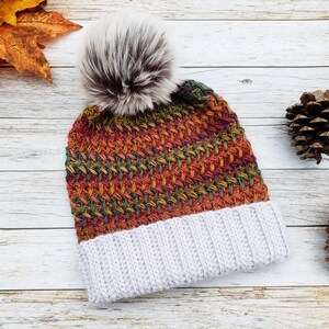 May include: A handmade knit beanie with a faux fur pom-pom. The hat features a woven pattern in shades of orange, red, green, and yellow, with a light gray ribbed brim. The pom-pom is white and brown.