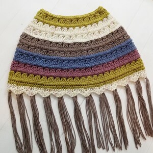 May include: A crocheted poncho with a multi-colored striped pattern in shades of brown, blue, pink, and green. The poncho has a scalloped edge and a fringe trim.