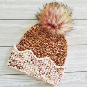 May include: A brown and cream crocheted beanie with a fluffy brown pom pom on top. The beanie has a scalloped edge.