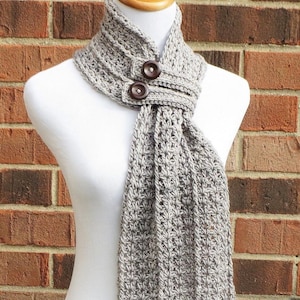 Puede incluir: Una bufanda de ganchillo gris con dos botones marrones. La bufanda está atada alrededor del cuello de un maniquí blanco.