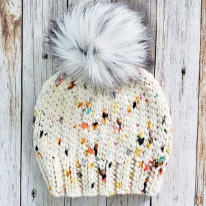 May include: A white knitted beanie with a fluffy white pom pom. The beanie has a speckled pattern with multi-coloured flecks.