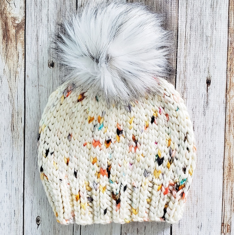 May include: A white knitted beanie with a fluffy white pom pom. The beanie has a speckled pattern with multi-coloured flecks.