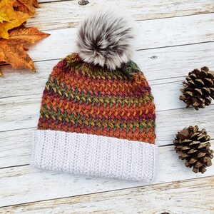 May include: A knitted beanie with a faux fur pom-pom. The hat features a color block design with a light gray ribbed brim and a woven pattern in shades of orange, green, and red. The pom-pom is a mix of gray and brown.