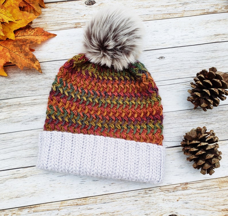 May include: A knitted beanie with a faux fur pom-pom. The hat features a color block design with a light gray ribbed brim and a woven pattern in shades of orange, green, and red. The pom-pom is a mix of gray and brown.