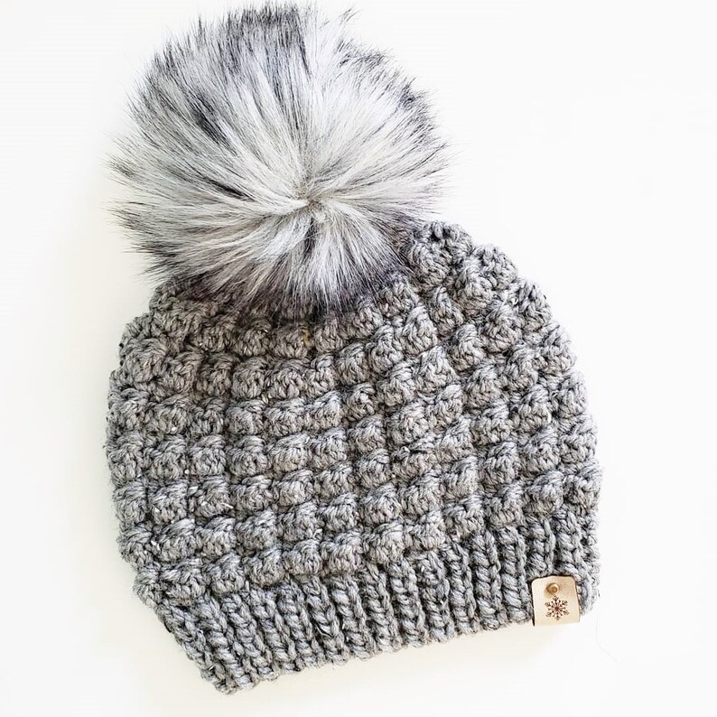 May include: Gray crocheted beanie with a fluffy white pom pom and a small brown leather tag with a snowflake design.