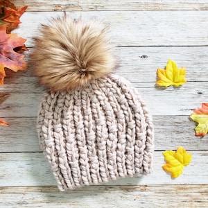 May include: A beige knitted beanie with a brown faux fur pom pom. The beanie is on a white wood background with fall leaves.