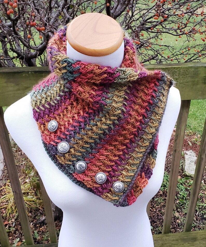 Op de afbeelding: Een gehaakte sjaal met een veelkleurig patroon van rood, oranje, groen en bruin. De sjaal heeft vijf zilveren knopen met een bloemmotief.