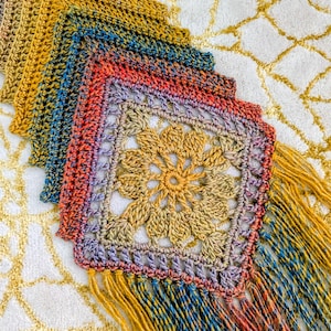 Könnte beinhalten: Handgefertigter Häkelschal in leuchtenden Farben. Der Schal zeigt ein quadratisches Muster mit einem zentralen Blumenmotiv, das durch Gelb-, Orange-, Blau- und Lilatöne verläuft. Er ist mit langen, gelben Fransen versehen.