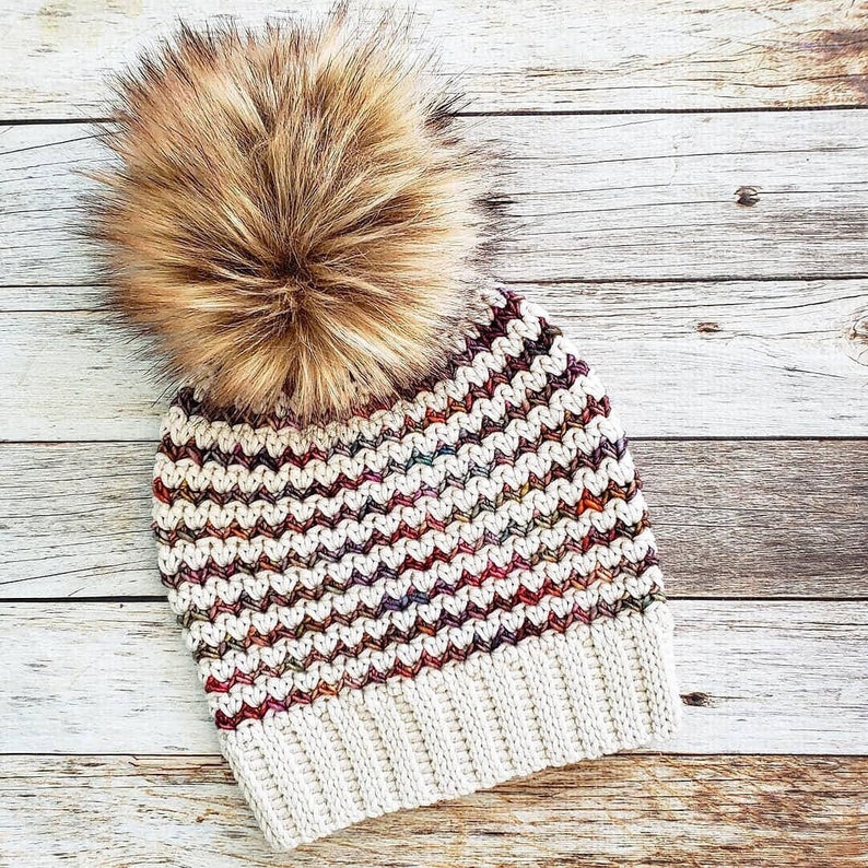 May include: A white crocheted beanie with a colorful chevron pattern and a brown faux fur pom pom.