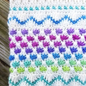 May include: A white crocheted blanket with a colorful geometric pattern. The pattern features rows of blue, green, purple, and pink stitches.
