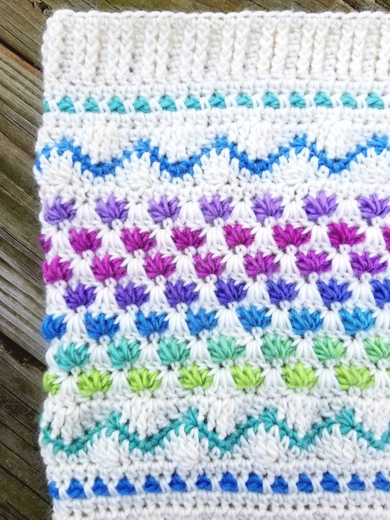 May include: A white crocheted blanket with a colorful geometric pattern. The pattern features rows of blue, green, purple, and pink stitches.
