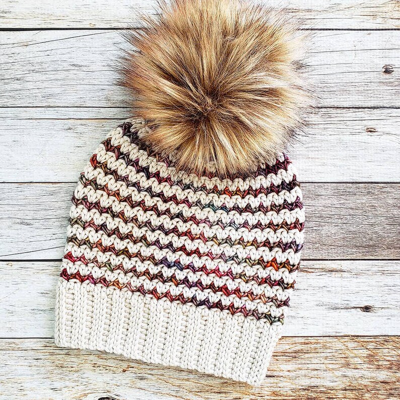 May include: A crocheted beanie with a brown faux fur pom pom. The beanie has a white base with a multi-colored stripe pattern.