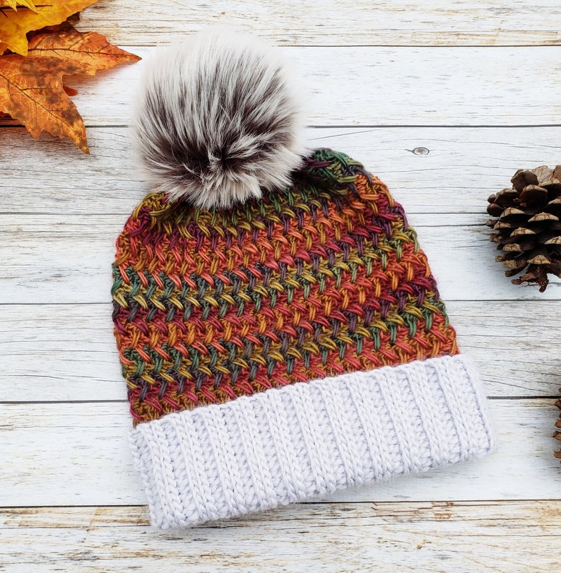 May include: A handmade knit beanie with a faux fur pom-pom. The hat features a woven pattern in shades of orange, red, green, and yellow, with a light gray ribbed brim. The pom-pom is white and brown.