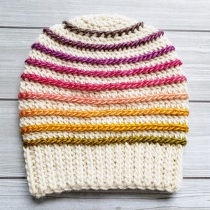 Pode incluir: Um gorro branco de croch&ecirc; com um padr&atilde;o de listras coloridas. As listras s&atilde;o em tons de marrom, roxo, rosa, laranja e verde.