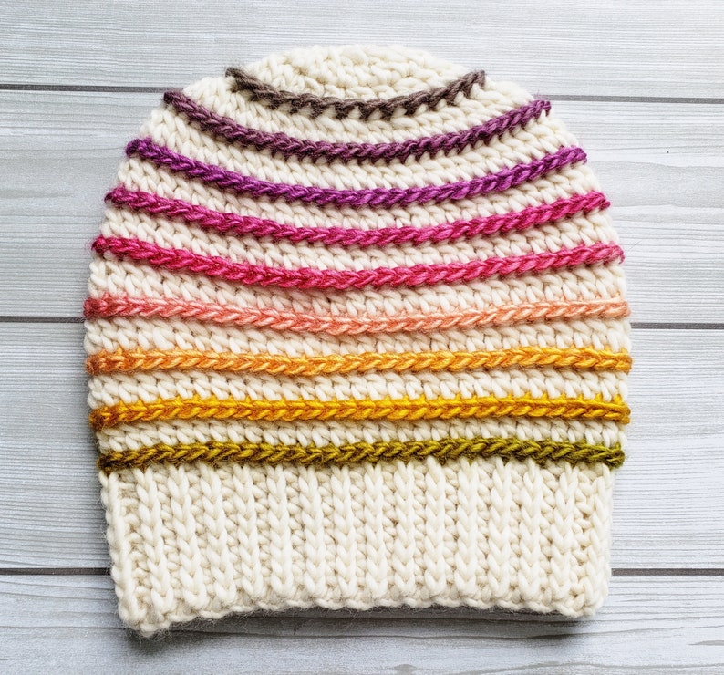 Pode incluir: Um gorro branco de croch&ecirc; com um padr&atilde;o de listras coloridas. As listras s&atilde;o em tons de marrom, roxo, rosa, laranja e verde.