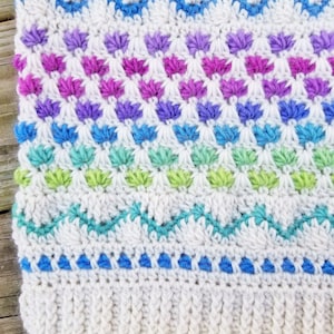 May include: A white crocheted blanket with a colorful geometric pattern. The pattern features rows of blue, green, purple, and pink stitches.