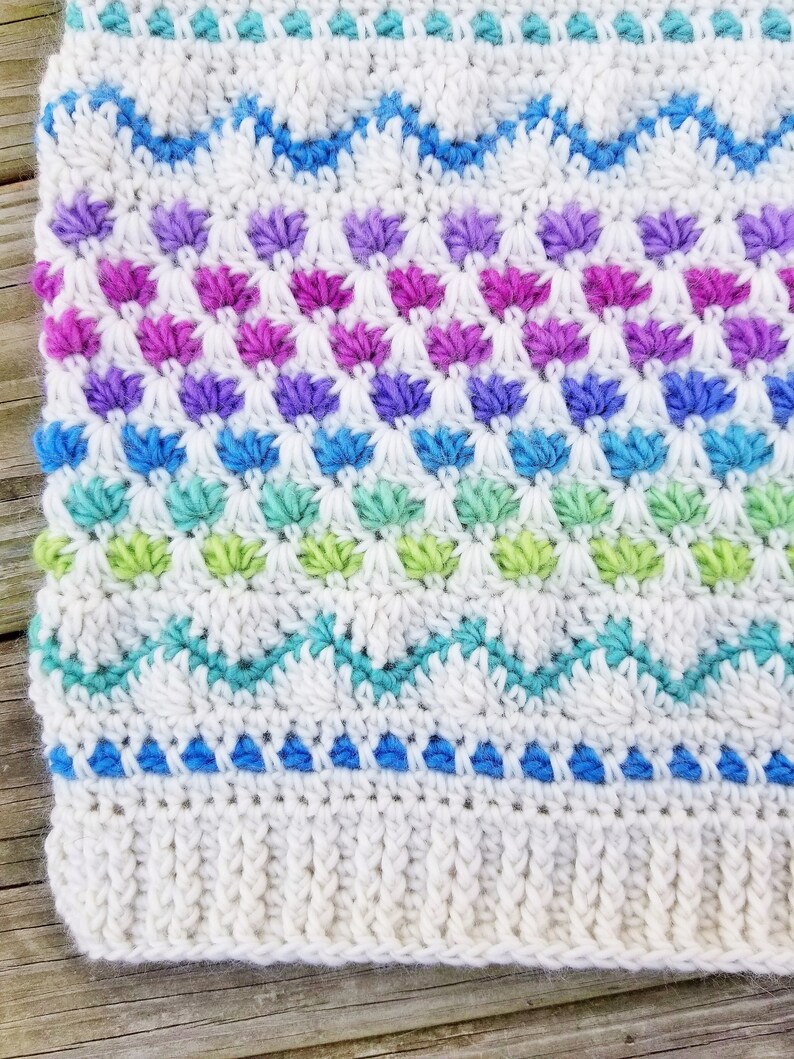 May include: A white crocheted blanket with a colorful geometric pattern. The pattern features rows of blue, green, purple, and pink stitches.