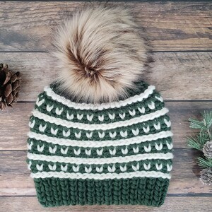 May include: A green and white knitted beanie with a faux fur pom pom. The beanie has a pattern of white stripes and green leaves.
