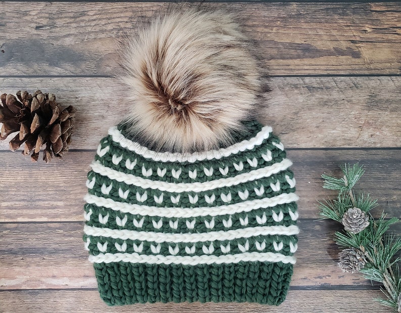 May include: A green and white knitted beanie with a faux fur pom pom. The beanie has a pattern of white stripes and green leaves.