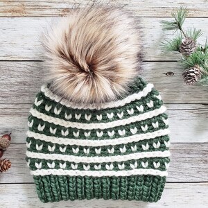 May include: A green and white crocheted beanie with a brown faux fur pom pom. The beanie has a striped pattern with white stripes and green leaves.
