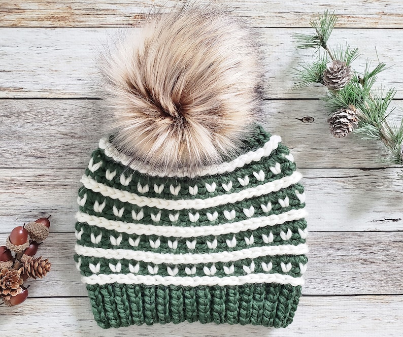 May include: A green and white crocheted beanie with a brown faux fur pom pom. The beanie has a striped pattern with white stripes and green leaves.