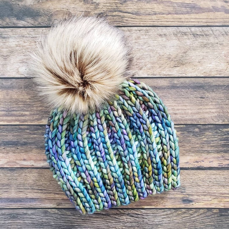 K&ouml;nnte beinhalten: Eine blaue, gr&uuml;ne und violette Strickm&uuml;tze mit einem flauschigen braunen Bommel.