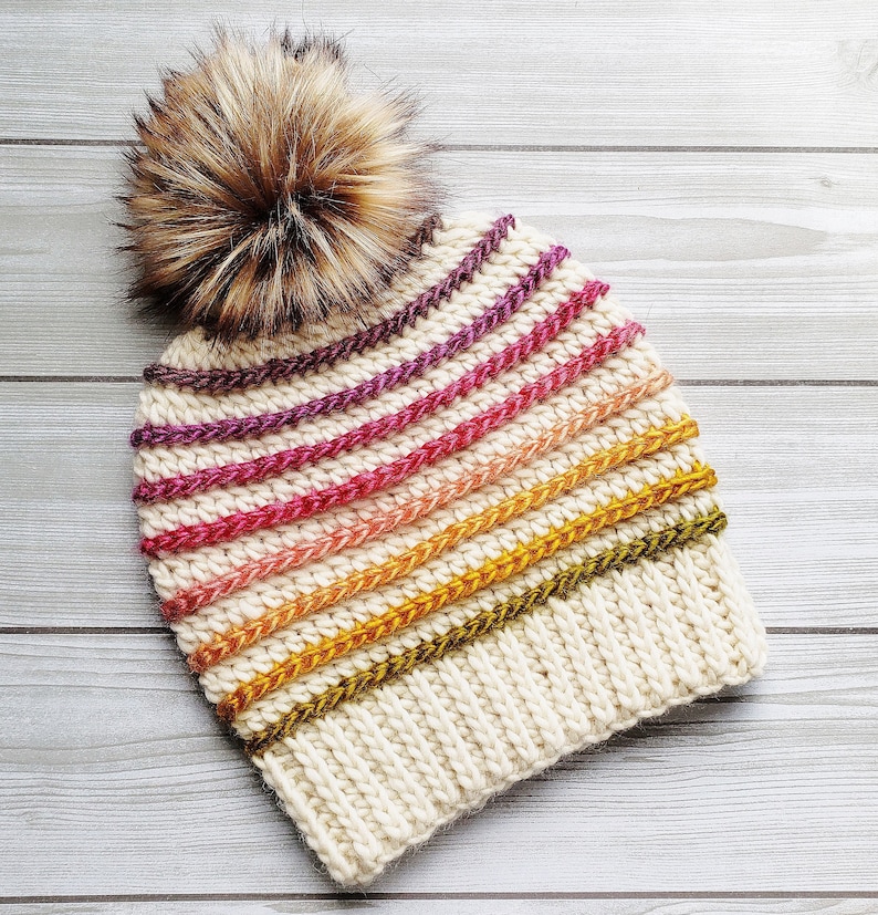 Pode incluir: Um gorro de malha branco com um padr&atilde;o listrado colorido em tons de rosa, laranja, amarelo e verde. O gorro tem um pompom marrom felpudo no topo.