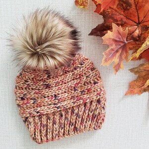 May include: A knitted beanie with a fluffy pom pom in shades of brown, orange, and pink. The beanie has a textured knit pattern and is made with a soft yarn.
