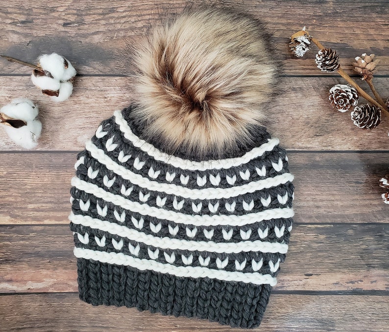 May include: A grey and white knitted beanie with a faux fur pom pom. The beanie has a striped pattern with white and grey yarn.