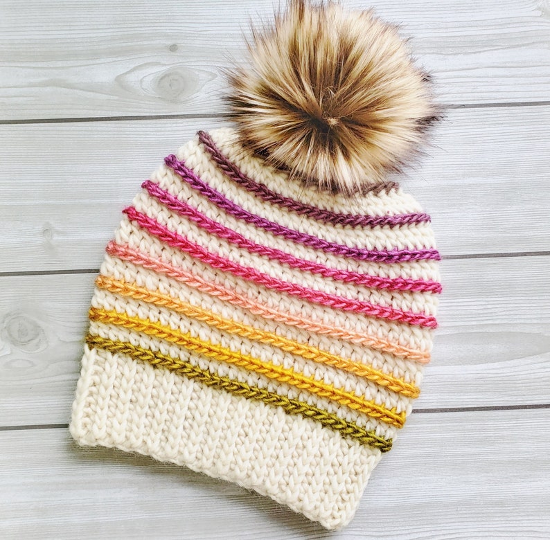 Pode incluir: Um gorro branco de croch&ecirc; com um padr&atilde;o listrado colorido em tons de rosa, laranja, amarelo e verde. O gorro tem um pompom de pele sint&eacute;tica marrom no topo.